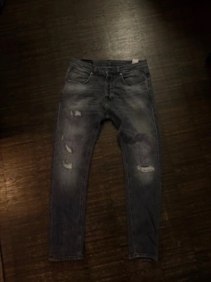 Dondup jeans - Säljer ett par grå jeans med slitningar och hål på låret och knät. Jeansen har fem fickor och klassisk knappgylf. Perfekta för en avslappnad och trendig look.