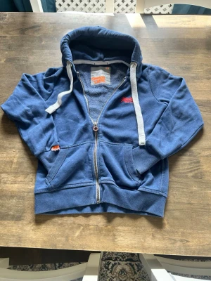 Blå SuperDry Zip-Up Tröja - Säljer en blå hoodie från Superdry med dragkedja, vita snören i huvan och fickor framtill. Hoodien har en liten orange Superdry-logga på ärmen och en röd logga på bröstet. Mjuk insida och ribbade muddar vid ärmslut och nederkant.