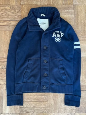 Marinblå kofta Abercrombie & Fitch - Marinblå kofta från Abercrombie & Fitch med broderad text 'PROP OF A&F 92' på bröstet och två vita ränder på ena ärmen. Tröjan har knäppning framtill, två fickor och ribbade muddar. Perfekt för en avslappnad och sportig stil.