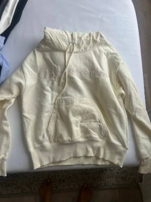 Vit hoodie från Champion - Säljer en vit hoodie från Champion med broderad logga på bröstet. Hoodien har en stor känguruficka framtill, ribbade muddar och dragsko i huvan. Perfekt för en chill och avslappnad stil.