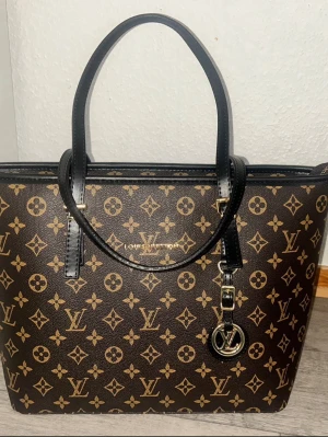 En stor handväska - Snygg handväska i klassiskt brunt monogrammönster. Väskan har svarta handtag, guldfärgade detaljer och en rund. Tillverkad i tåligt canvasmaterial med läderdetaljer. Perfekt för dig som gillar lyxig och tidlös stil.