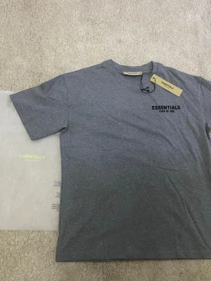 Grå t-shirt Essentials Fear of God - Snygg grå t-shirt från Essentials Fear of God med svart logga tryckt på bröstet. Klassisk rund hals och korta ärmar. Perfekt basplagg med stilren design som passar till det mesta. Tillverkad i mjuk bomull för skön komfort.