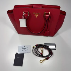 Röd handväska från Prada - Snygg röd handväska från Prada i strukturerat läder med guldfärgade metalldetaljer. Väskan har både handtag och avtagbar axelrem samt logotyp framtill. Medföljer äkthetskort och dustbag. Perfekt för dig som vill ha en lyxig accessoar. 0688