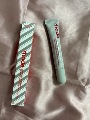 Rhode Peptide Lip Treatment Peppermint - Peppermint Peptide Lip Treatment från Rhode i en stilren tub med röd text. Läppbalsamet har en ljusgrå förpackning och kommer i en matchande randig ask. Perfekt för dig som vill ha återfuktade och mjuka läppar med en fräsch mintig känsla.
