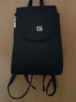 Svart mini ryggsäck - Liten svart mini ryggsäck. Okänt märke. Fint skick. En stor ficka och två små fickor; en i det större last utrummet och en på baksidan av väskan. Det finns dragkedjor på sidan av väskan för att kunna öka utrymmet till max kapacitet. Gjord i Thailand.