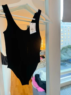 Svart ribbad body med breda axelband - Snygg svart body med breda axelband och rundad halsringning. Tillverkad i ett ribbat stretchigt material som sitter tight och bekvämt. 