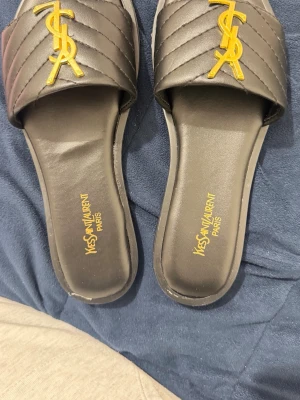Svarta  sandaler med gulddetalj - Säljer ett par svarta sandaler Sandalerna har öppen tå och platt sula, tillverkade i konstläder med lyxig känsla. Perfekta för en trendig och stilren look. Helt nya 