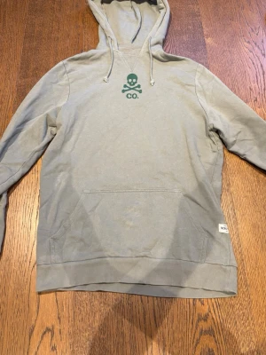 Ljusgrå hoodie med döskalletryck - Ljusgrå hoodie med huva och dragsko, samt en stor magficka framtill. På bröstet finns ett grönt tryck med döskalle och texten 'CO.'. Tillverkad i mjukt bomullsmaterial och har ribbade muddar vid ärmslut och nederkant. Perfekt för en avslappnad stil.