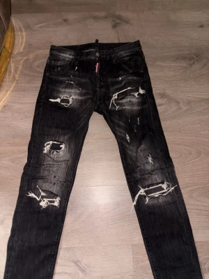 Svarta Dsquared2 jeans  - Säljer ett par svarta Dsquared2 jeans med kraftiga slitningar och hål på både fram- och baksida. Jeansen har en tvättad look och detaljerade slitningar vid knän och lår. Perfekta för dig som gillar en edgy och trendig stil.