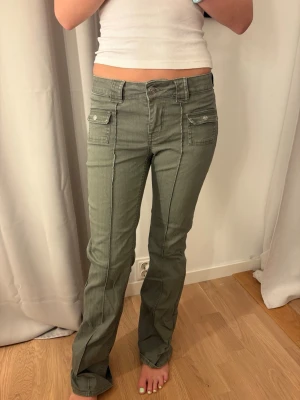 Gröna bootcut jeans med fickor lågmidjade khaki - Snygga gröna bootcut jeans med markerade sömmar och två stora fickor framtill med lock och knapp. Jeansen har hög midja och stängs med knapp och dragkedja. Perfekta för en trendig och avslappnad stil.