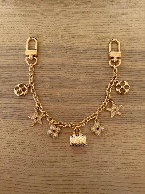  Vuitton guldfärgad väsksmycke - Lyxigt guldfärgat väsksmycke från  Vuitton med flera hängande berlocker i form av stjärnor, blommor och en liten väska. Smycket har två karbinhakar för enkel fastsättning på väskan och är tillverkat i metall med glittrande detaljer.