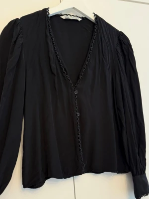 Blus med volangkant - Stilren och elegant svart blus från Zara. Har en smickrande V-ringning, knäppning framtill samt söta ögle- och spetsdetaljer längs kanterna och runt ärmsluten. Tillverkad i ett mjukt och fallande material med långa ärmar.