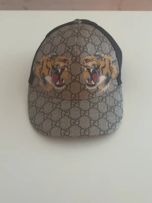 Gucci keps tiger  - ÄKTA!!!                                                                  Snygg keps från Gucci med klassiskt GG-mönster i beige och brunt. Framtill finns två detaljerade tigertryck i gult och svart med röda inslag. Baksidan är i svart mesh för extra ventilation och justerbar passform. Perfekt för dig som vill sticka ut.