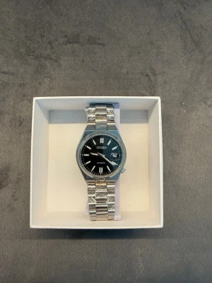 Citizen silverfärgad klocka - Stilren klocka från Citizen med silverfärgad länk i metall och svart urtavla. Klockan har tydliga vita markeringar, datumvisning och automatisk rörelse. Perfekt accessoar för dig som gillar klassisk och tidlös design.