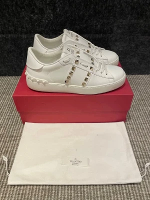 Vita Valentino Rockstud sneakers - Säljer ett par vita Valentino Rockstud sneakers i läder med ikoniska guldfärgade nitar längs sidorna och på plösen. Klassisk låg modell med vita snören och diskret logga på plösen. Kommer med originalpåse och röd box. Köpte dem nya på Plick men de var tyvärr försmå. Skriv för frågor. De är endast testade.