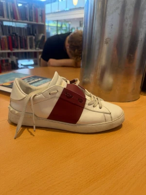 Vit sneaker med vinröd detalj - Säljer en stilren vit sneaker med en bred vinröd rem över snörningen. Skon har vita snören, låg profil och nitar på hälen. Tillverkad i slätt läder och passar perfekt till streetwear eller casual outfits.