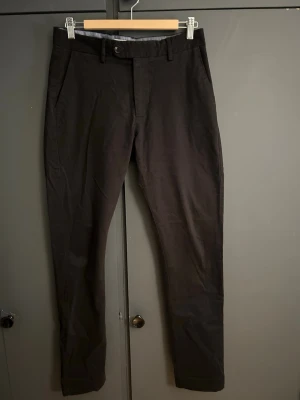 Svarta chinos från John Henric - Snygga svarta chinos från John Henric med smal passform och klassisk design. Byxorna har två sidofickor fram, två bakfickor med knapp och bälteshällor. Tillverkade i ett mjukt bomullsmaterial som ger en clean look. Super chysta och ger dig verkligen en stilig look. 