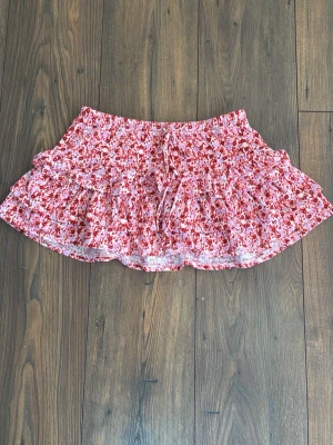 Blommig volangkjol från Gina Tricot - Supersöt kort kjol från Gina Tricot med blommigt mönster i rosa och rött. Kjolen har volanger och en liten snörning framtill. Perfekt för varma dagar och ger en riktigt somrig vibe.Ordinarie pris - ca 300kr