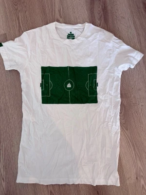 Vit t-shirt med grön fotbollsplan - Vit t-shirt i bomull med ett stort grönt tryck av en fotbollsplan på bröstet. Klassisk rund hals och korta ärmar. Perfekt för dig som gillar sportig stil och vill sticka ut med en unik design.