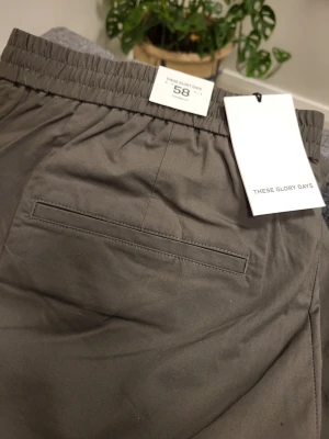 Gråa mjukisbyxor These Glory Days - Snygga olivgröna mjukisbyxor från These Glory Days med elastisk midja och raka ben. Byxorna har en bakficka och sidofickor, tillverkade i ett mjukt och tunt material som känns skönt mot huden. Perfekta för en avslappnad och stilren look.