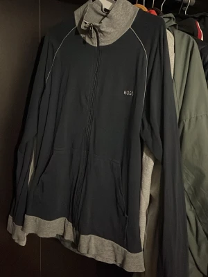 Mörkblå ziptröja från BOSS - Snygg mörkblå hoodie från BOSS med dragkedja framtill och gråa detaljer vid krage och nederkant. Hoodien har diskret logga på bröstet och två fickor fram. Perfekt för en avslappnad och stilren look.