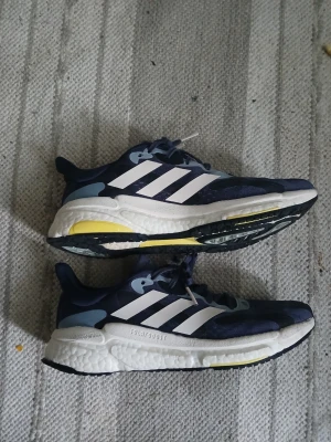 Adidas Solarglide Boost sneakers strlk 40.5 - Mörkblå Adidas Solarglide Boost sneakers med vita och ljusblå detaljer samt gula inslag på sulan. Skorna har klassiska tre vita ränder på sidan, mesh-ovandel för bra ventilation och en tjock, stötdämpande Boost-sula. Perfekta för dig som gillar sportig stil. Storlek 40.5 de är i mkt bra skick.  Använda endast ett fåtal ggr 
