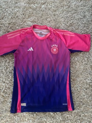 Tysklands matchtröja rosa - Snygg Tyskland matchtröja från Adidas i rosa och lila med zigzag-mönster. Tröjan har korta ärmar, rund hals och vita Adidas-loggor samt Tysklands landslagsemblem med fyra stjärnor på bröstet. Tillverkad i funktionsmaterial för sport.