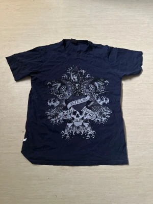 Ed hardy t-shirt - Säljer min Ed hardy t-shirt med stort grafiskt tryck framtill som visar dödskalle, banderoll och fiskar. Texten 'Love Kills Slowly' syns i motivet. T-shirten har korta ärmar och rund hals. Perfekt för dig som gillar edgy streetstyle.
