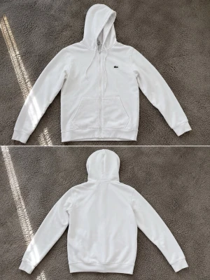 Vit hoodie med dragkedja från Lacoste - Snygg vit hoodie från Lacoste med dragkedja framtill och klassisk krokodil-logga på bröstet. Hoodien har huva med dragsko och två stora fickor framtill. Perfekt för en clean och stilren look.