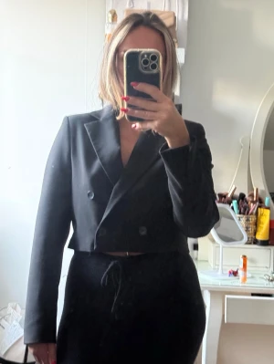 Svart cropped kavaj med dubbelknäppning - Snygg svart cropped kavaj med dubbelknäppning och klassisk krage. Kavajen har en rak passform och långa ärmar, perfekt för en trendig och stilren look. Materialet ser ut att vara en mjuk och slät textil som ger en elegant känsla.