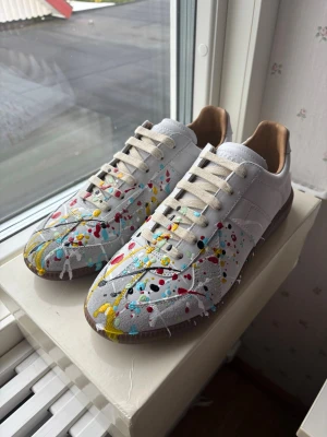 Maison margiella Paint splatter  - Helt oanvända unika vita sneakers med färgstänk i gult, rött, blått och svart. Skorna har låga skaft, beige snörning och bruna sulor. Ovandelen är i läder och textil, vilket ger en cool och konstnärlig vibe. Perfekta för dig som vill sticka ut med din stil.