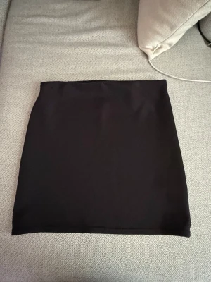 Svart enkel kortkjol - En stilren svart kortkjol med rak passform. Kjolen är enfärgad och har en minimalistisk look som passar till många olika outfits. Materialet känns mjukt och stretchigt, vilket gör den bekväm att bära. Perfekt att matcha med en crop top eller oversized tröja.
