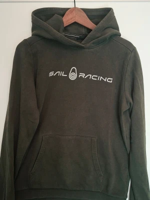 Olivgrön hoodie från Sail Racing - Säljer en olivgrön hoodie från Sail Racing med vit logga och text på bröstet. Hoodien har en stor ficka framtill och en huva med liten logga bak. Tillverkad i mjukt bomullsmaterial och har långa ärmar. Perfekt för en avslappnad och sportig stil.