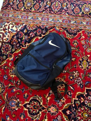 Mörkblå ryggsäck från Nike - Snygg mörkblå ryggsäck från Nike med vit logga. Ryggsäcken har flera fack med dragkedja, vadderade axelband och detaljer i svart. Perfekt för skola, träning eller utflykter. Tillverkad i slitstarkt syntetmaterial.
