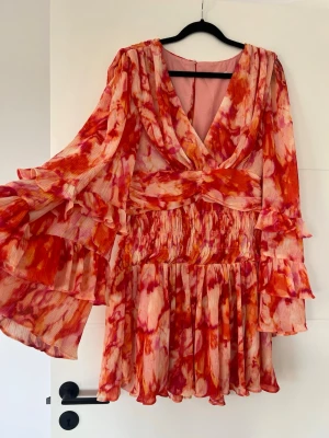 NA-KD x Sanna Jörnvik - Färgstark kortklänning med blommigt mönster i rött, rosa och orange. Klänningen har v-ringning, markerad midja med twistad detalj och vida volangärmar. Tillverkad i ett lätt, plisserat material som ger ett flowy intryck. Perfekt för sommarens alla tillfällen.