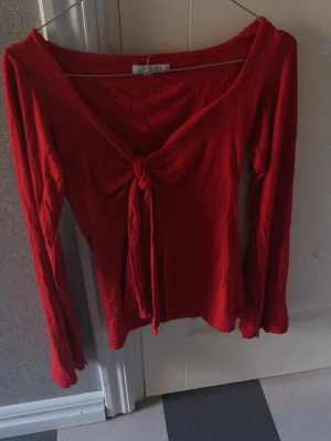 Röd långärmad topp med knyt - Snygg röd långärmad topp med djup v-ringning och knyt framtill. Toppen har en figurnära passform och är tillverkad i mjukt bomullstyg. Perfekt för dig som vill ha en enkel men ändå trendig look.