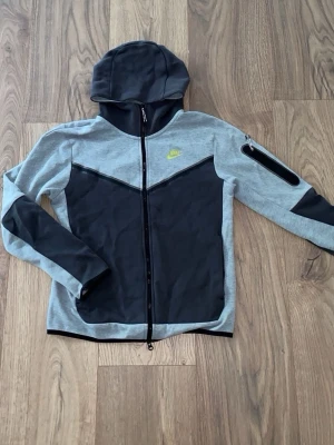 Nike tech fleece (S) - Snygg hoodie från Nike i grått och svart med dragkedja framtill och huva. Har en cool detalj med ficka på ärmen och limegrön Nike-logga på bröstet. Perfekt för dig som gillar sportig och avslappnad stil.