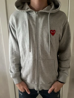 Grå hoodie med rött hjärta - Säljer en grå zip-hoodie med huva och dragsnören. På bröstet finns ett litet rött hjärta med ögon, vilket ger en unik och lekfull touch. Hoodien har fickor framtill och är tillverkad i mjukt sweatshirtmaterial. Perfekt för en avslappnad och trendig stil. 