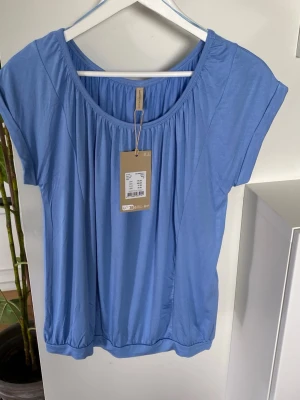 Säljer en ljusblå t-shirt/tröja/topp från SoyaConcept Ny med prislapp  - Säljer en ljusblå t-shirt/tröja/topp från Soyaconcept av modellen SC-Marica med fint veckad halsringning och en elastisk resår i midjan.  Tyget är mjukt!   Rund halsringning med veckdetalj Korta ärmar med upprullad detalj Lyocell- och elastanblandning  Material: 93% lyocell, 7% elastan Maskintvätt i högst 30 °C. Ska ej torktumlas Strykning, högsta temperatur 110 °C.    