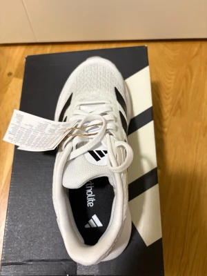 Vita Adidas sneakers med svarta ränder - Snygga vita sneakers från Adidas med klassiska svarta ränder på sidan. Ovandelen är i meshmaterial för bra andningsförmåga och skon har en bekväm sula. Perfekta för dig som gillar sportig och stilren look.
