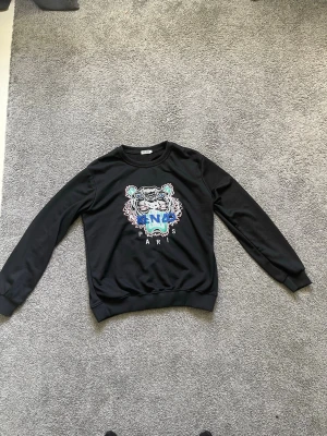Svart Kenzo sweatshirt - Svart sweatshirt från Kenzo med ikoniskt broderat tigermotiv i flera färger och texten 'KENZO PARIS' på bröstet. Tröjan har rund halsringning och ribbade muddar vid ärmslut och nederkant. Perfekt statement-plagg för dig som gillar streetwear.