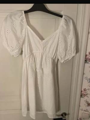 Vit kort klänning med puffärm - Söt vit kort klänning med broderade blommor och puffiga kortärmar. Klänningen har v-ringning och är gjord i bomull med fina spetsdetaljer. Perfekt för sommarens alla tillfällen och ger en romantisk vibe.