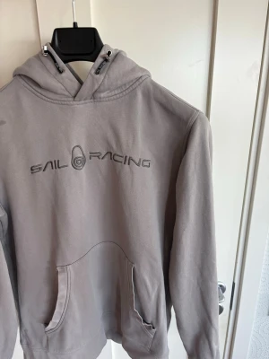 Grå hoodie från Sail Racing - Snygg grå hoodie från Sail Racing med stor logga broderad på bröstet. Hoodien har en klassisk känguruficka framtill och justerbar huva med snören. Perfekt för en avslappnad och sportig stil.