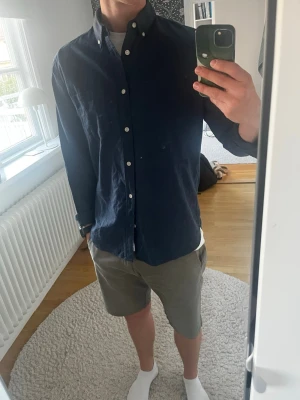 Mörkblå linneskjorta med knappar - Säljer en stilren mörkblå skjorta i linne med klassisk krage och vita knappar. Skjortan har lång ärm och en bröstficka. Perfekt för dig som gillar en enkel men snygg look. Typ aldrig använt den, köpte den för 499