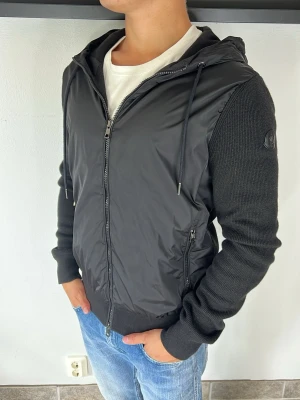 Moncler cardigan  - Snygg svart hoodie med dragkedja framtill och huva med dragsko. Framsidan är i glansigt material medan ärmar och rygg är ribbstickade. Två fickor med dragkedja på sidorna och diskret patch på ärmen. Perfekt till jeans för en avslappnad look.