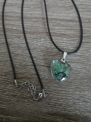 Halsband med hjärthänge och blågrön blomma - Unikt halsband med svart läderrem och ett hjärtformat hänge i transparent glas. Inuti hänget finns en liten blågrön blomma och en fjäril som ger en naturlig och cool look. Perfekt för dig som gillar naturinspirerade accessoarer och vill sticka ut.