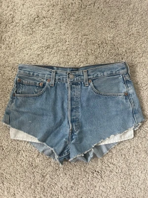 Blå jeansshorts levis - Säljer ett par klassiska blå jeansshorts med fransig kant från levis. Shortsen har knäppning med knappar framtill. Perfekta för sommaren och ger en avslappnad vibe. Finns vissa defekter på ena fickan som man kan se på bilden! Skriv om du undrar något!