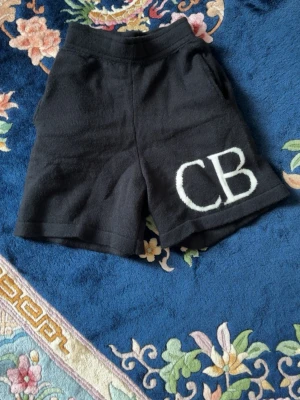 CB shorts - Svarta stickade shorts med bred resår i midjan och stora vita bokstäver 'CB' på ena benet. Shortsen har två fickor bak och en mjuk, lite loose passform. Perfekta för en chill och avslappnad stil.