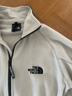 Vit fleecejacka från The North Face - Mysig vit fleecejacka från The North Face med dragkedja framtill och hög krage. Jackan har logga broderad på bröstet och nacken samt svarta detaljer längs dragkedjan och nederkanten. Perfekt för alla möjliga tilfällen!👌