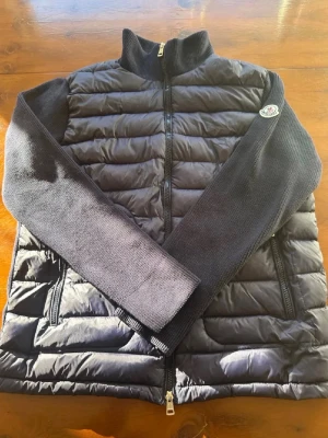 Moncler Cardigan  - Snygg mörkblå cardigan från Moncler med stickade ärmar och quiltad front. Jackan har hög krage, dragkedja framtill och två fickor med dragkedja. Moncler-logga på ärmen och innerfoder med serietidningsmönster. Perfekt för kalla vinterdagar. NFC SCAN FUNKAR!